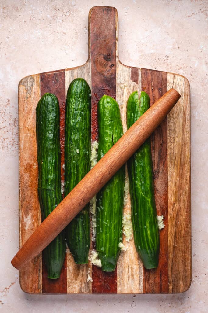Cucumbers being smashed with a wooden rolling pin on a cutting board