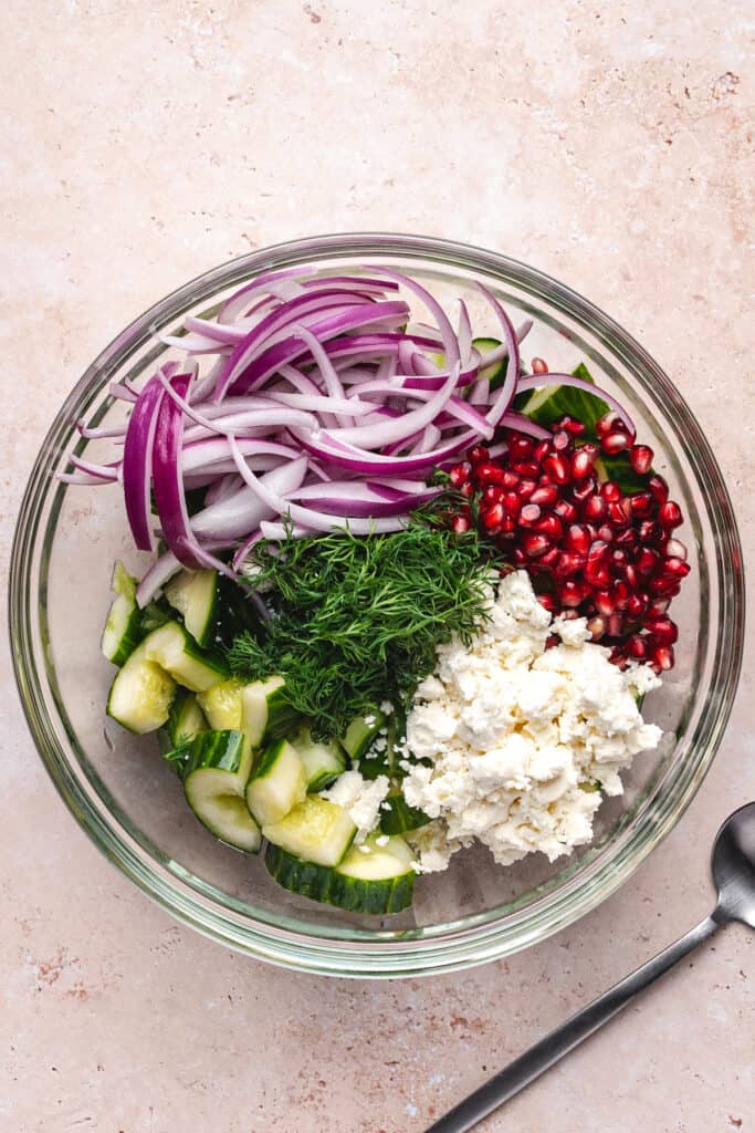 Bowl with unmixed salad ingredients: cucumber, red onion, dill, feta, and pomegranate seeds