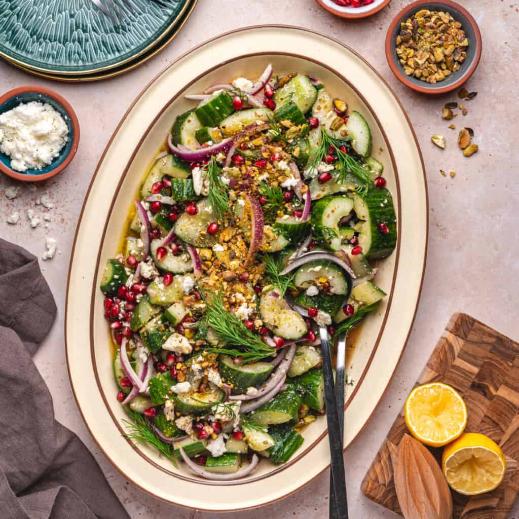 Overhead square image of smashed cucumber salad in a large platter, surrounded by ingredients and serving tools