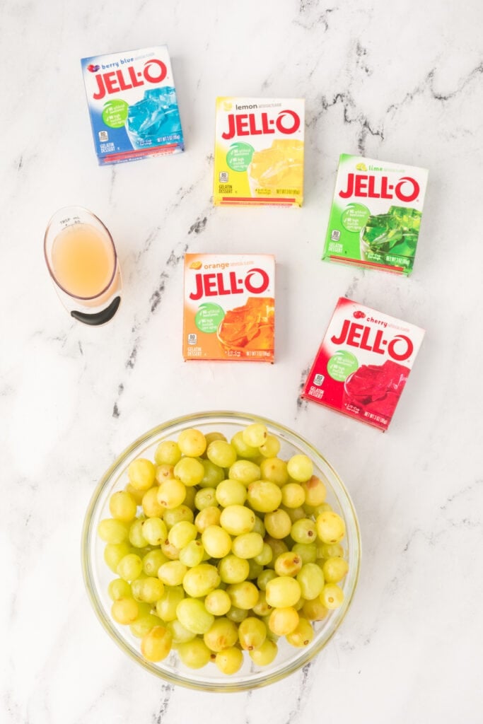 Ingredients for Sour Patch Grapes, including green grapes, lemon juice, and five boxes of colorful Jello mix on a white background