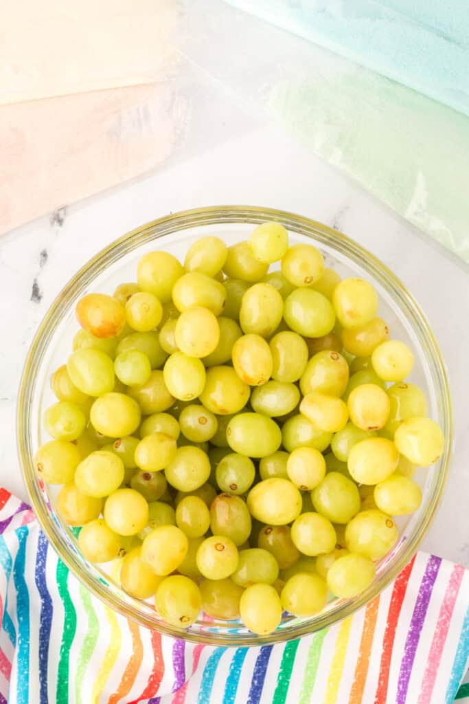 Bowl of rinsed green grapes surrounded by bags of colored gelatin powder and a rainbow-striped cloth