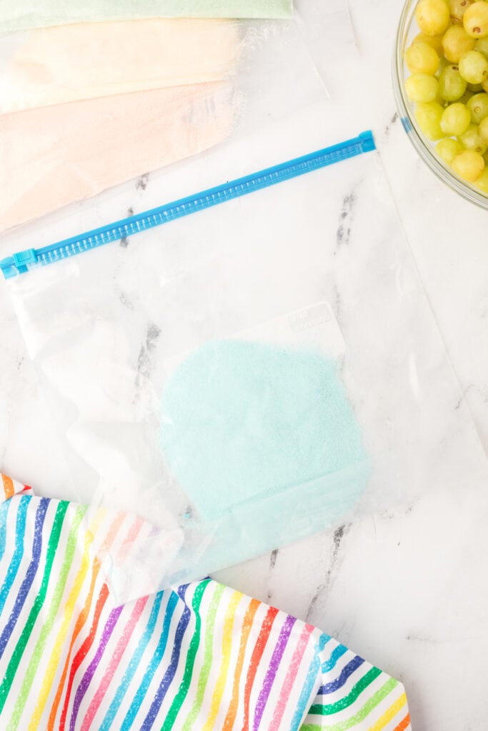 Close-up of a zip-top bag filled with blue Jello powder, next to other pastel powders and a bowl of grapes