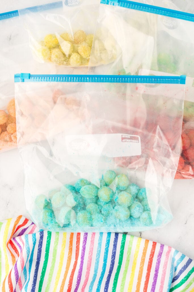Several zip-top bags filled with different colored gelatin-coated grapes resting on a rainbow cloth