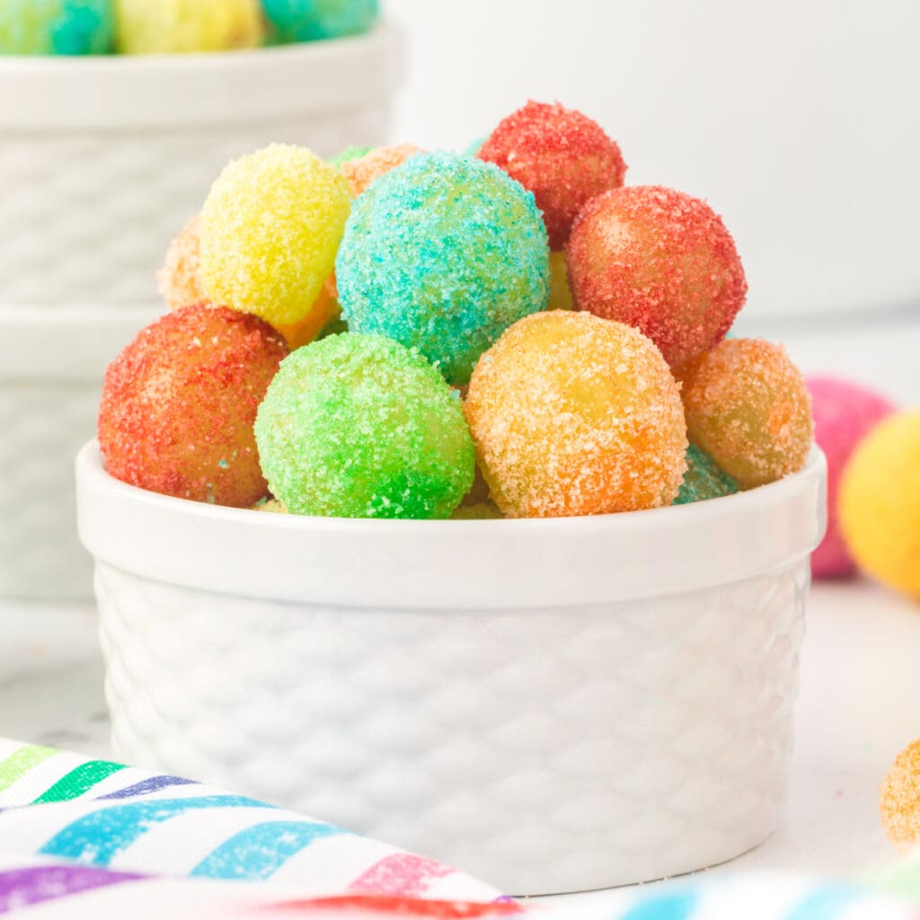 Square crop of a white ramekin filled with vibrant Sour Patch Grapes on a striped rainbow napkin