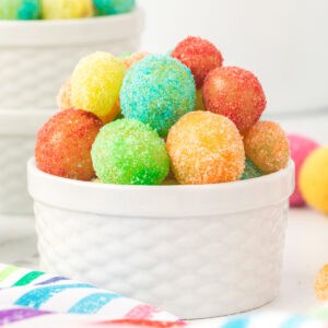 Square crop of a white ramekin filled with vibrant Sour Patch Grapes on a striped rainbow napkin