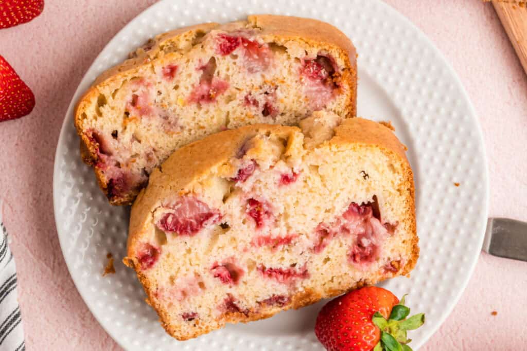 Two thick slices of strawberry bread on a white plate with a fresh strawberry beside them