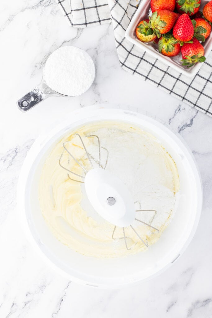Powdered sugar added to creamed butter in a mixing bowl for frosting.