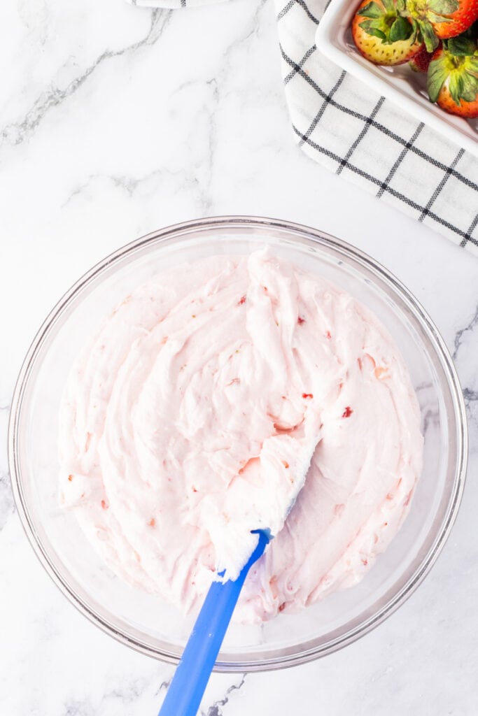 Creamy pink strawberry frosting with small strawberry pieces in a clear bowl and spatula.