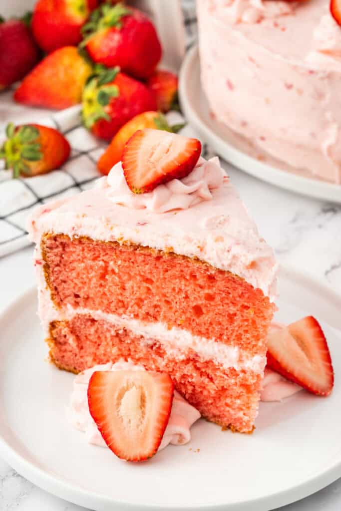 Slice of strawberry cake with pink frosting and sliced strawberries on top, served on a white plate.