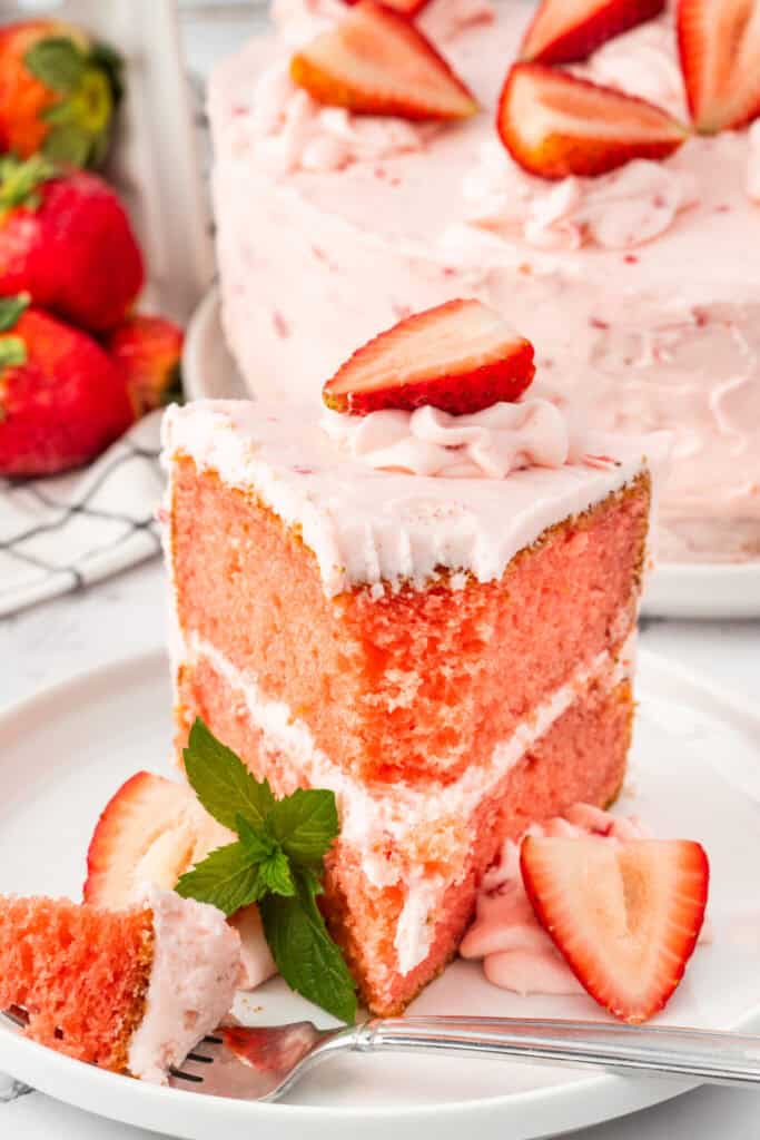 Forkful of strawberry cake with a mint garnish, served beside a full slice and decorated with fresh strawberries.