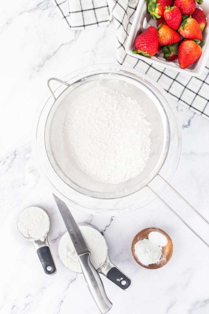 Sifting cake flour into a bowl surrounded by measuring cups and fresh strawberries.