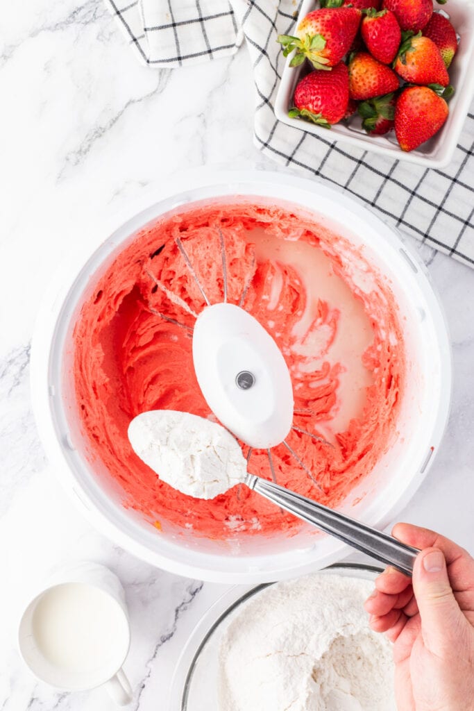 Flour being added to pink batter with a splash of buttermilk in the mixing bowl.