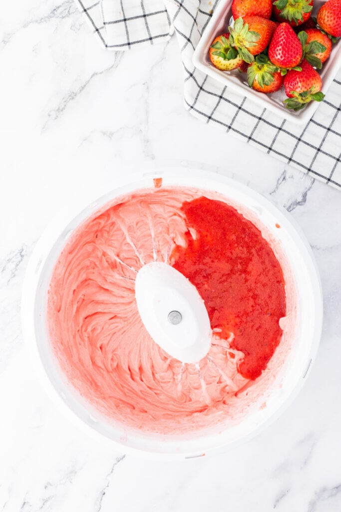 Strawberry purée being added to the pink cake batter in a large white mixing bowl.