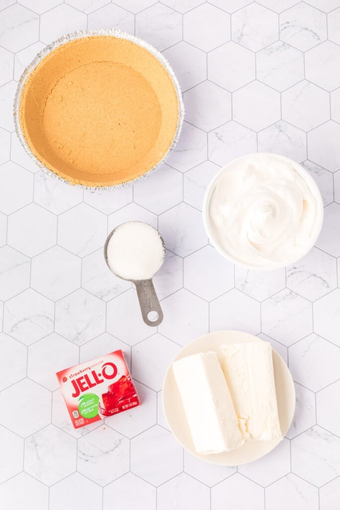 Overhead view of cream cheese, whipped topping, sugar, strawberry Jello, and graham cracker crust on a white hex tile surface