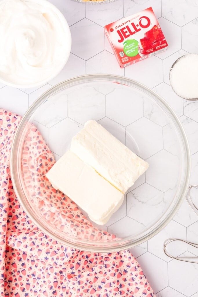 Cream cheese blocks in a glass bowl with ingredients for strawberry Jello pie arranged around it