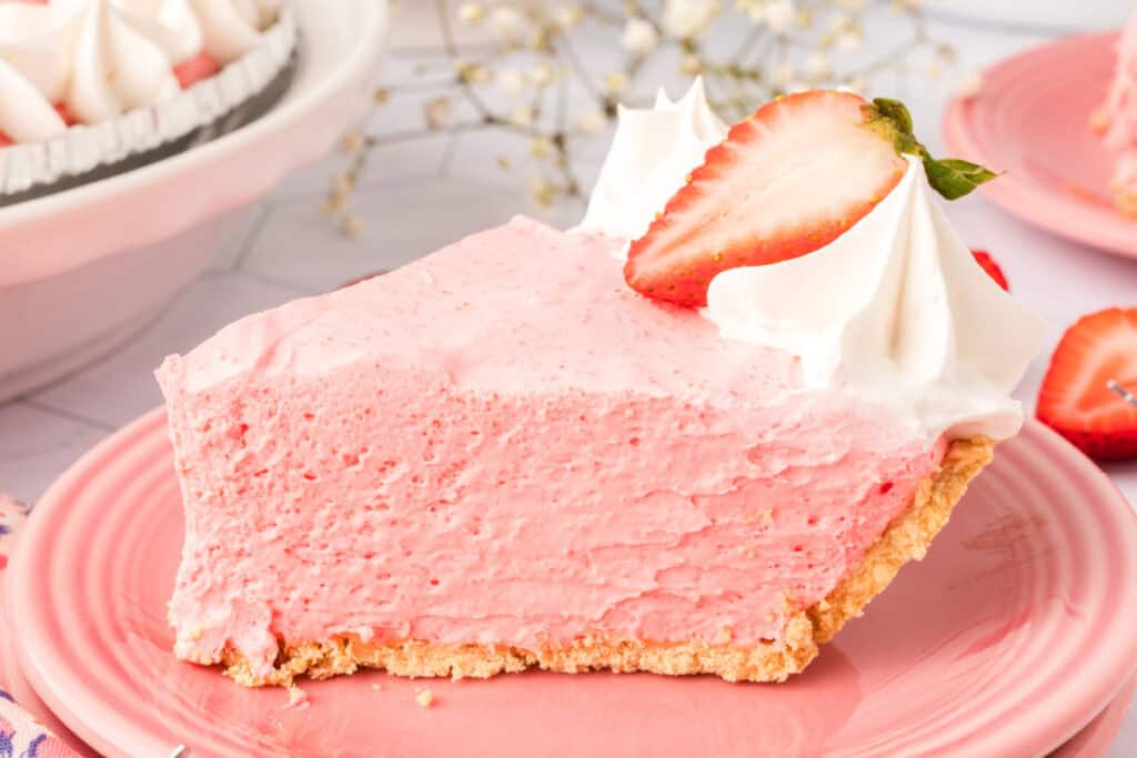 Close-up of a slice of strawberry cream pie on a pink plate, topped with whipped cream and a strawberry half