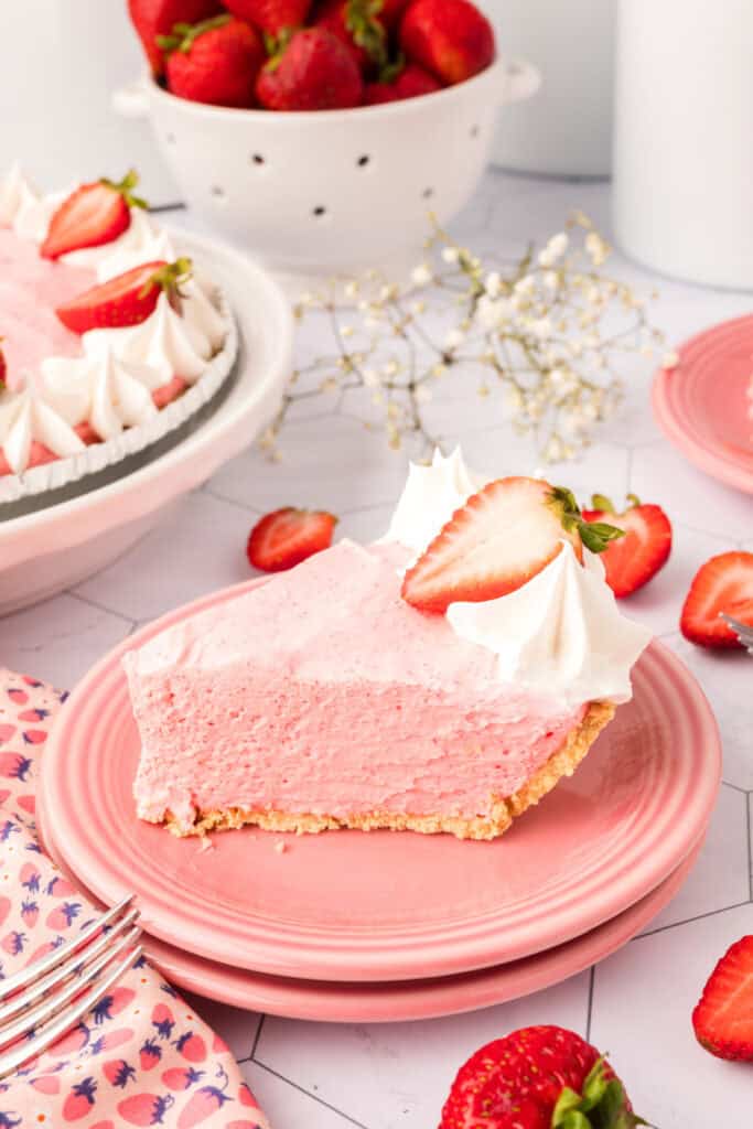 Overhead shot of a plated slice of strawberry cream pie with whipped cream and a strawberry, with the full pie in the background