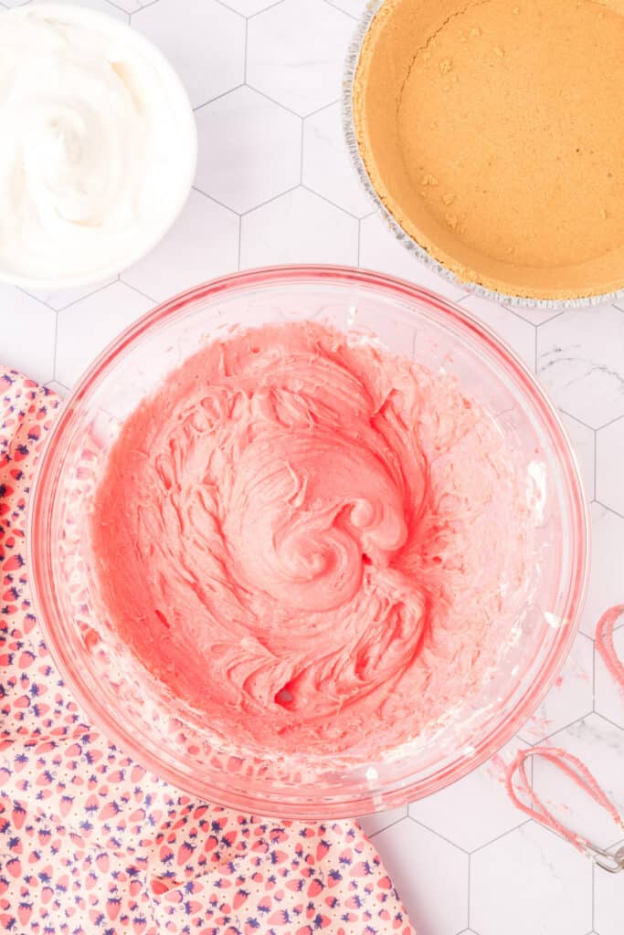 Strawberry filling fully mixed and bright pink in a large glass bowl, graham crust off to the side