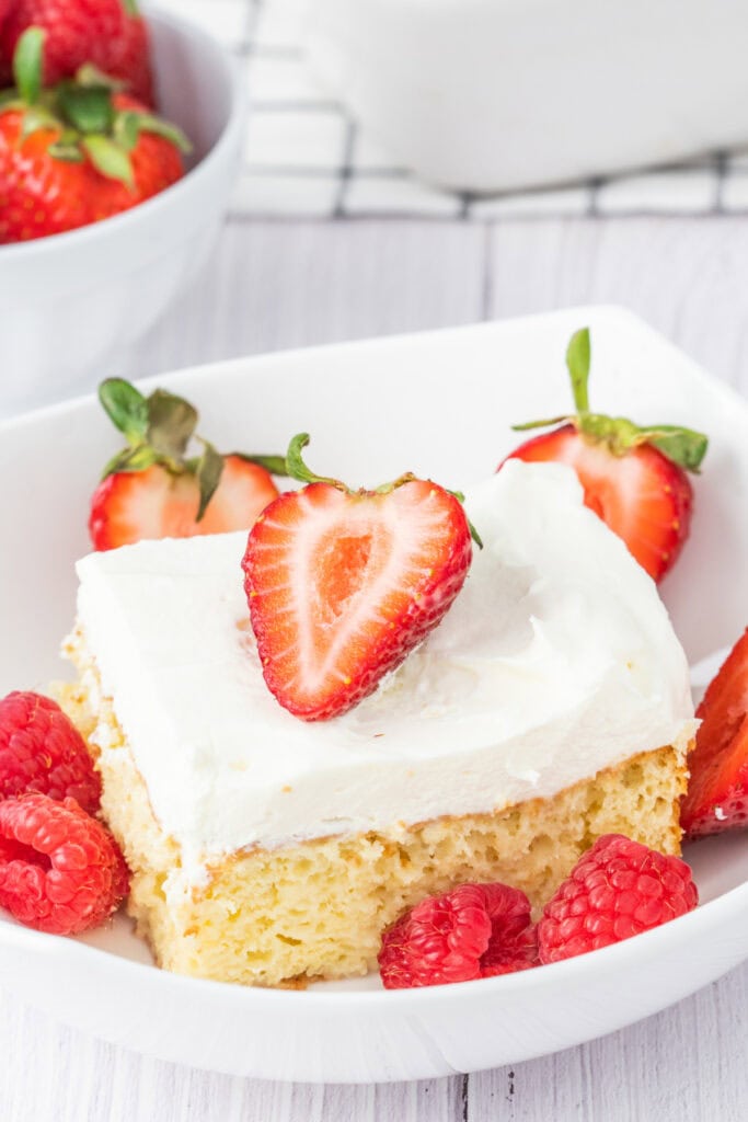 Closeup of a slice of tres leches cake topped with whipped cream, a halved strawberry, and surrounded by fresh raspberries in a white bowl.
