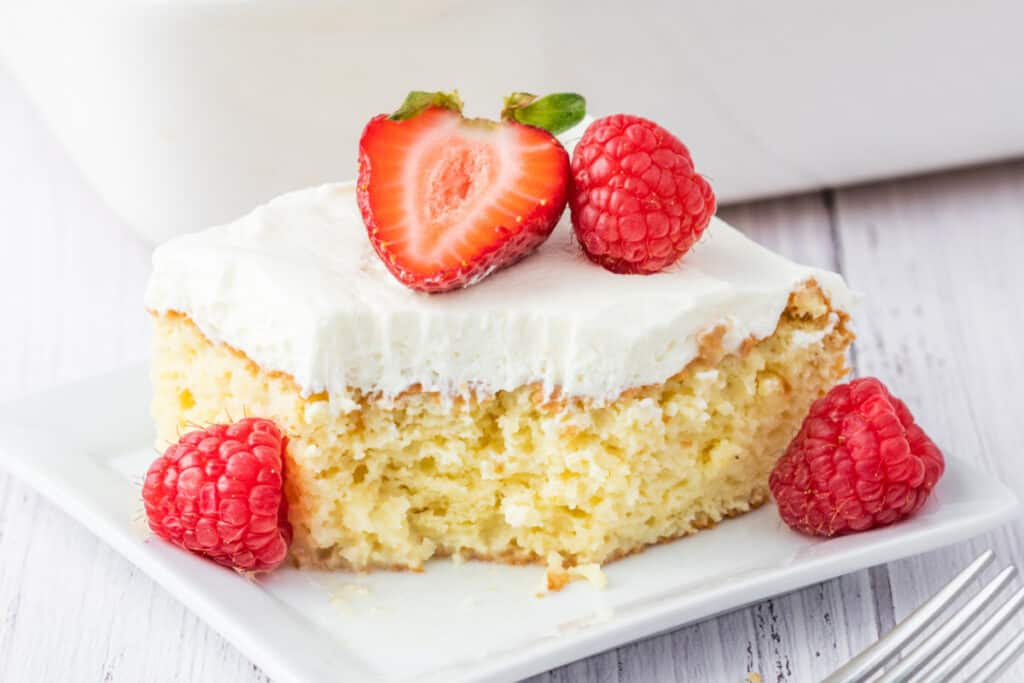 Slice of tres leches cake with whipped cream frosting, garnished with a halved strawberry and raspberries, served on a white square plate.