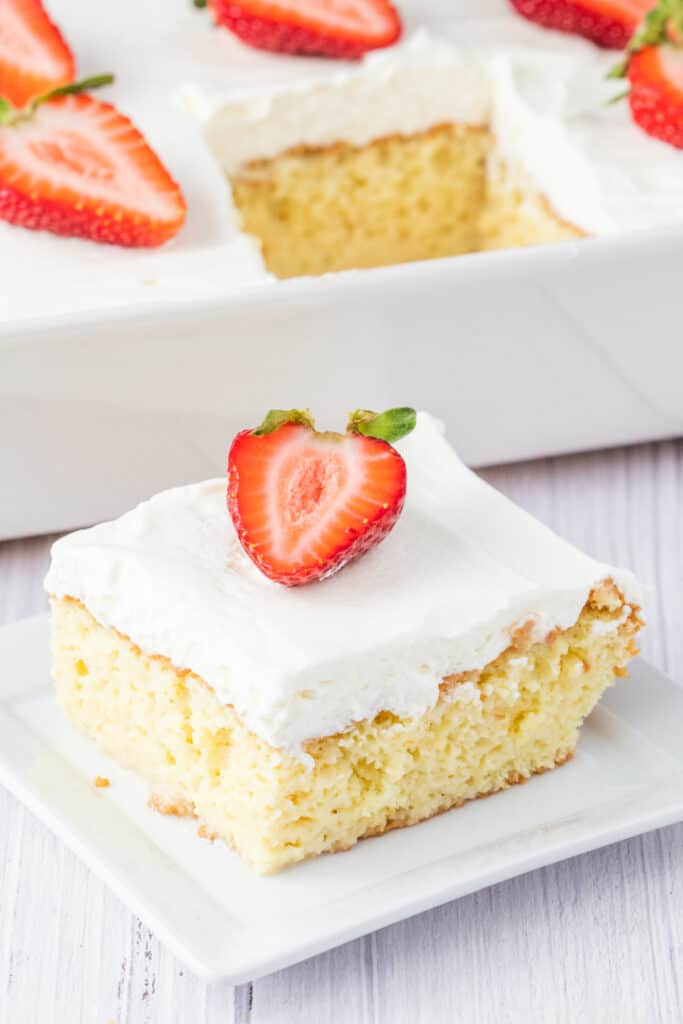 Single square of tres leches cake on a plate, topped with whipped cream and a fresh strawberry, with more cake visible in the background pan.