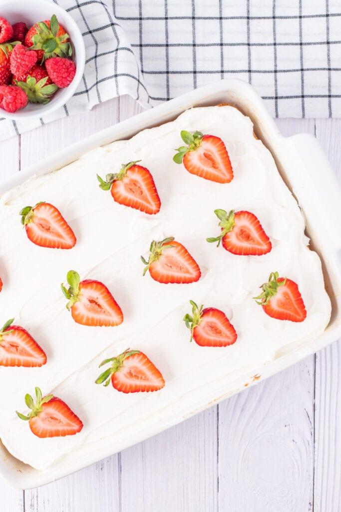 Overhead view of a full pan of tres leches cake topped with whipped cream and halved strawberries, with a bowl of fresh berries nearby.