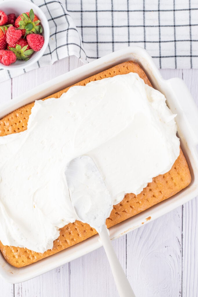 Frosting being spread over a poked tres leches cake in a white baking dish, with a bowl of raspberries and strawberries on the side.