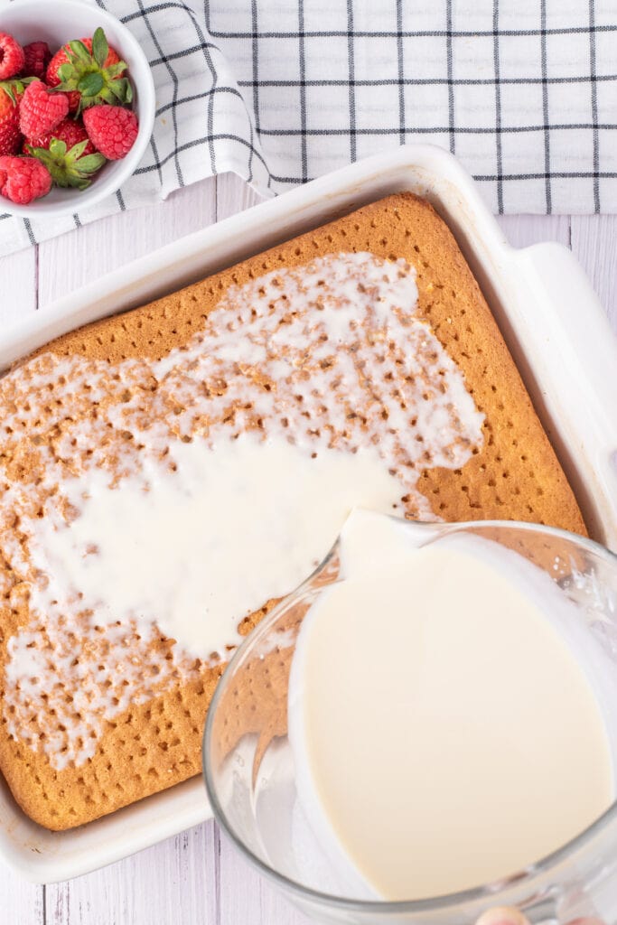 Tres leches milk mixture being poured over the poked cake in a white dish, ready for soaking, next to a bowl of fresh berries.