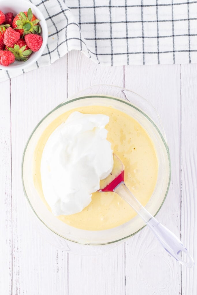 Whipped egg whites being folded into cake batter in a glass bowl with a red spatula, part of the cake mixing process.
