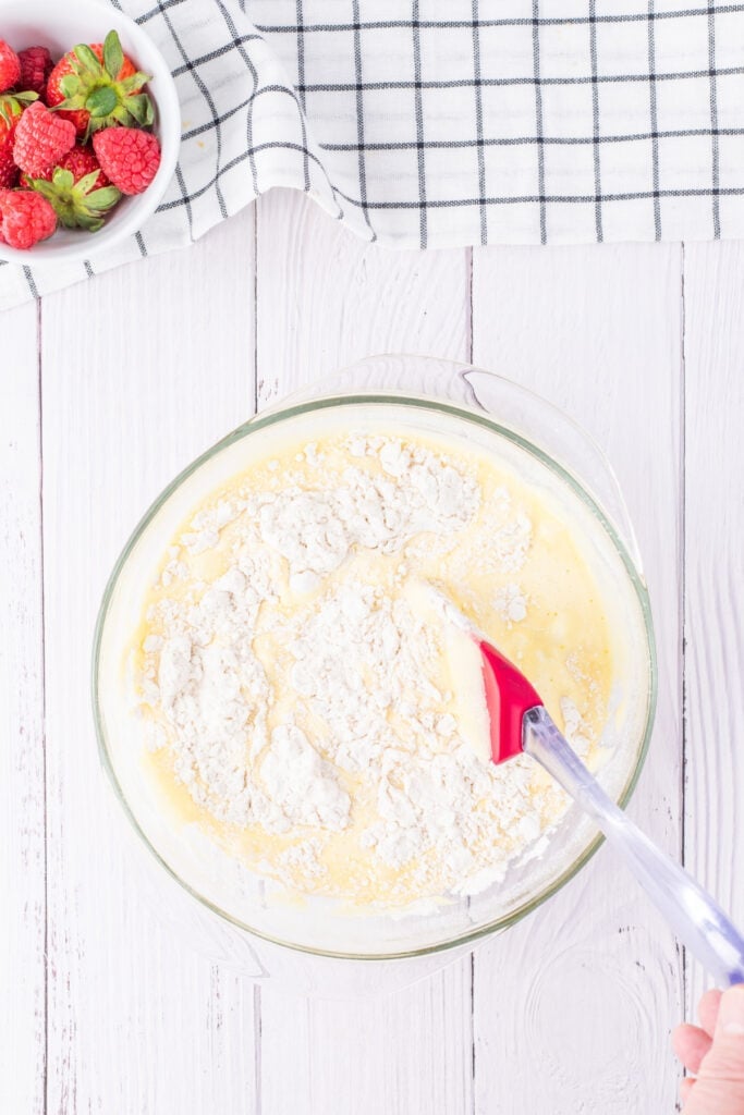 Dry ingredients being folded into egg yolk mixture in a glass bowl with a red spatula, part of the batter preparation for tres leches cake.