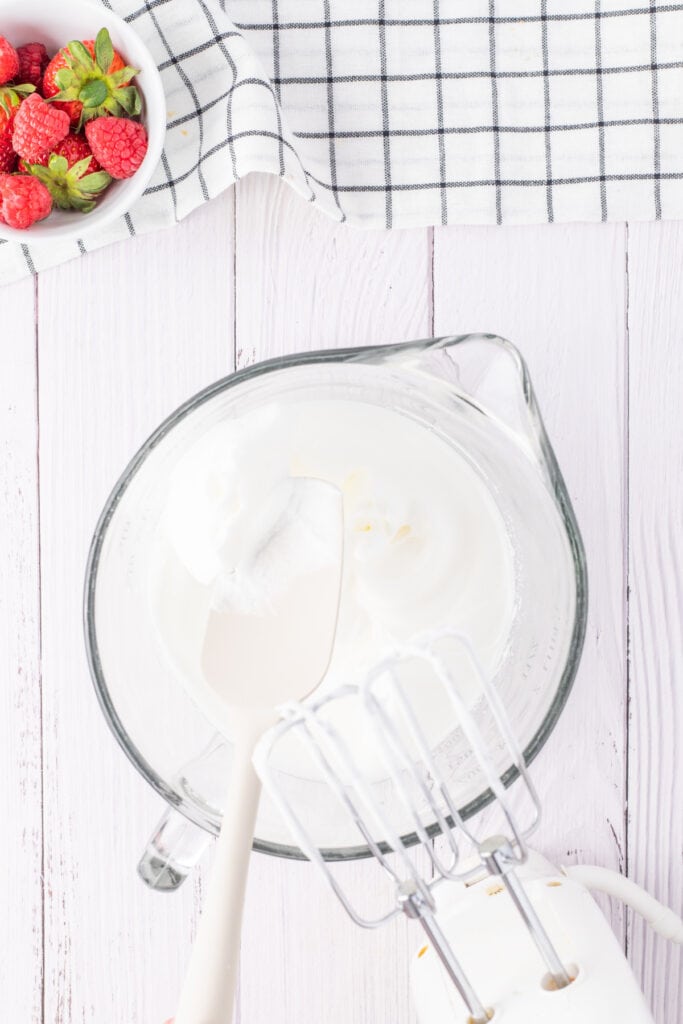 Whipping cream being mixed in a glass bowl with a spatula and beaters, preparing the whipped topping for tres leches cake.