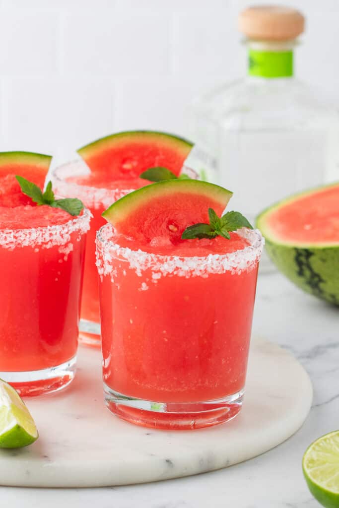 Three glasses of watermelon margaritas on a white surface with salt rims and watermelon slices