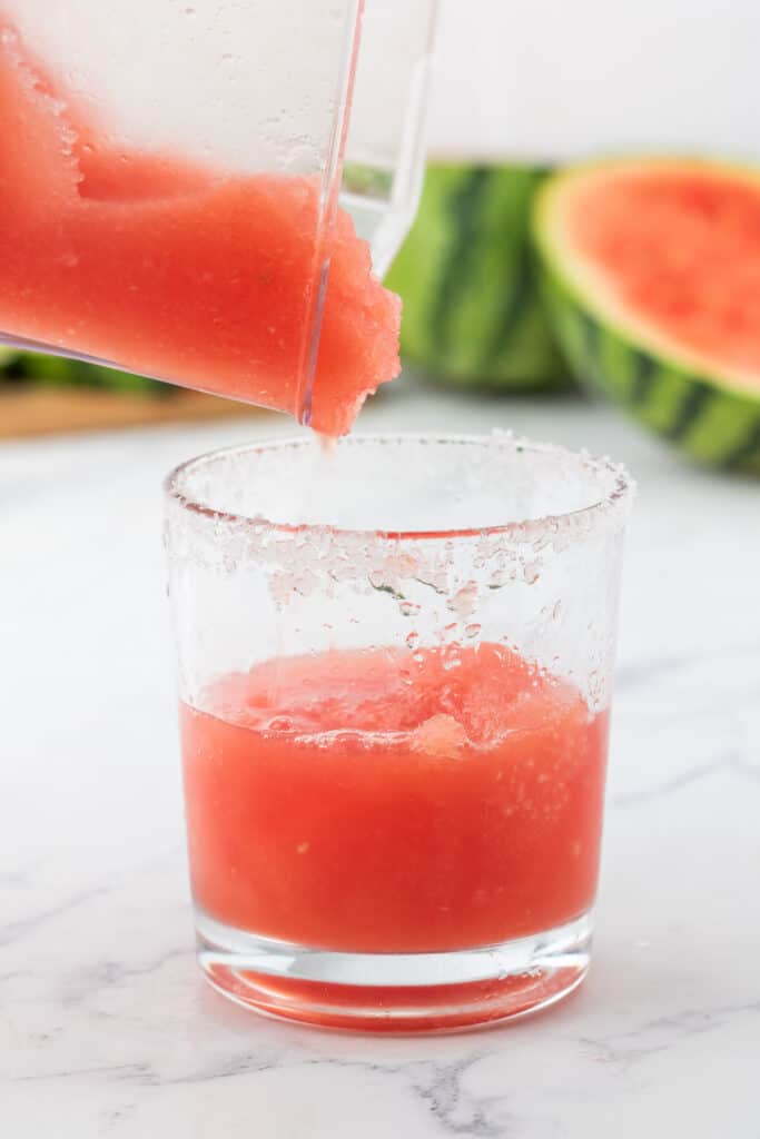 Blended watermelon margarita being poured from a blender into a salt-rimmed glass