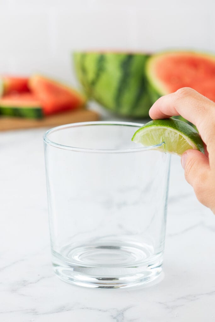 Hand rubbing a lime wedge along the rim of a clear glass with watermelon in the background