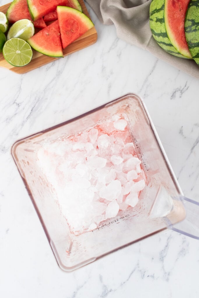 Ice added to a blender with watermelon and lime in the background on a white surface