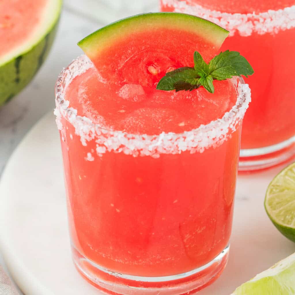 Square image of a bright red frozen watermelon margarita with a salted rim, garnished with a watermelon wedge and mint