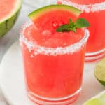 Square image of a bright red frozen watermelon margarita with a salted rim, garnished with a watermelon wedge and mint