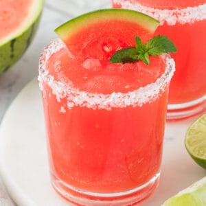 Square image of a bright red frozen watermelon margarita with a salted rim, garnished with a watermelon wedge and mint