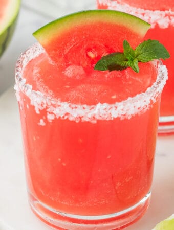 Square image of a bright red frozen watermelon margarita with a salted rim, garnished with a watermelon wedge and mint