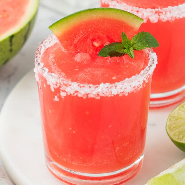 Square image of a bright red frozen watermelon margarita with a salted rim, garnished with a watermelon wedge and mint
