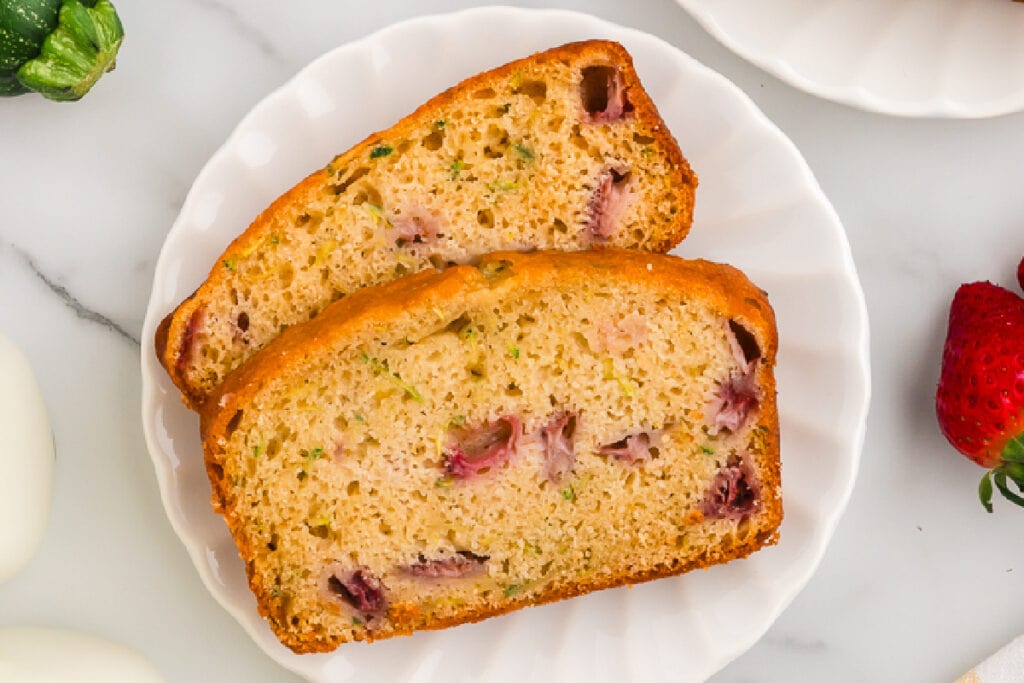 Overhead view of two slices of strawberry zucchini bread on a white plate with bits of fruit and zucchini visible