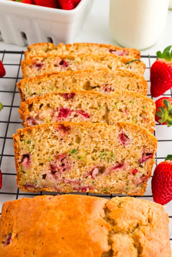 Sliced strawberry zucchini bread on a cooling rack surrounded by fresh strawberries and a bottle of milk