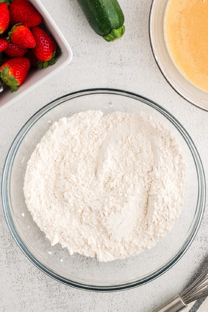 Dry ingredients fully mixed in a glass bowl next to fresh strawberries and zucchini