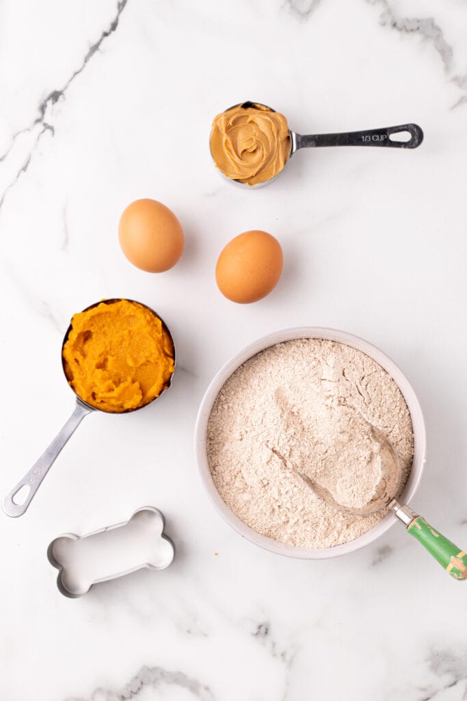 Ingredients for homemade dog treats arranged on a marble surface with a bone-shaped cookie cutter.