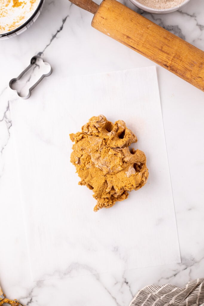Ball of pumpkin dog treat dough placed on parchment paper, ready to be rolled out, with a bone-shaped cookie cutter and vintage rolling pin nearby.