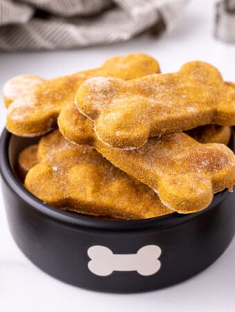 Square image of pumpkin dog treats stacked in a black bowl with bone design.