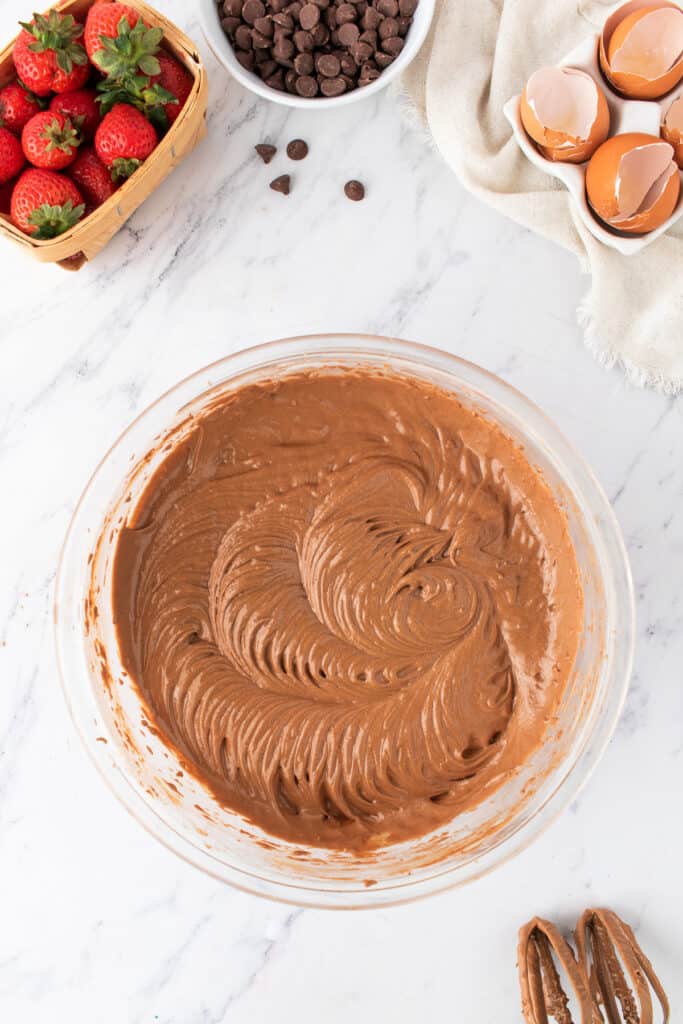 Bowl of chocolate cake batter with smooth, swirled texture, surrounded by fresh strawberries and chocolate chips.