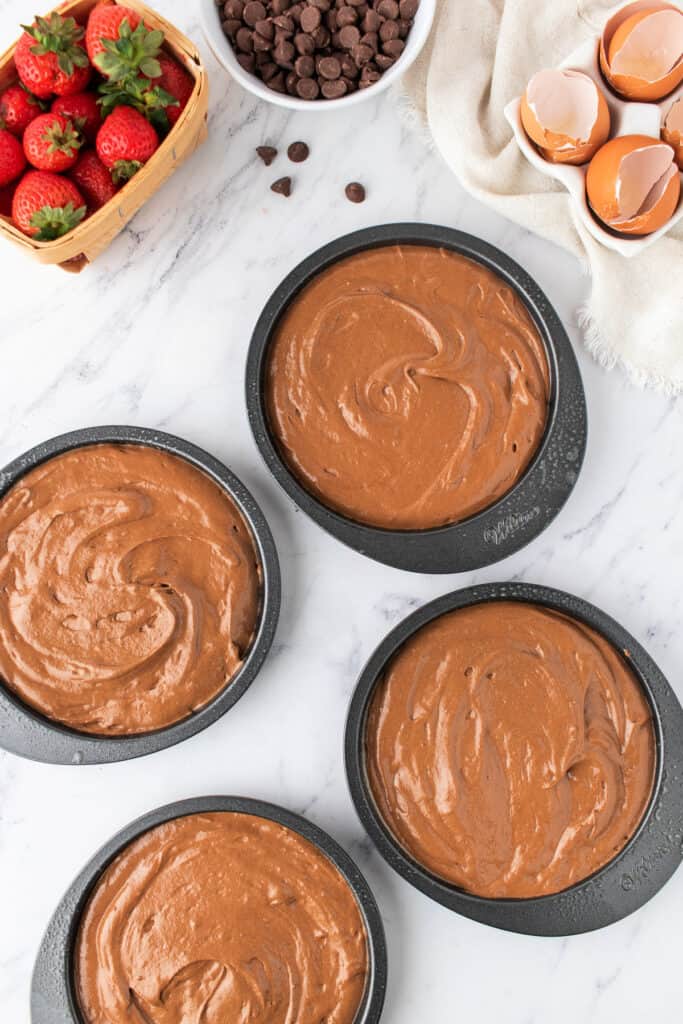 Four cake pans filled with chocolate batter, ready for baking, with strawberries and chocolate chips nearby.