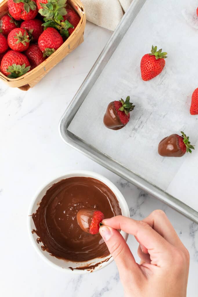 Hand dipping a fresh strawberry into melted chocolate over a parchment-lined baking sheet.