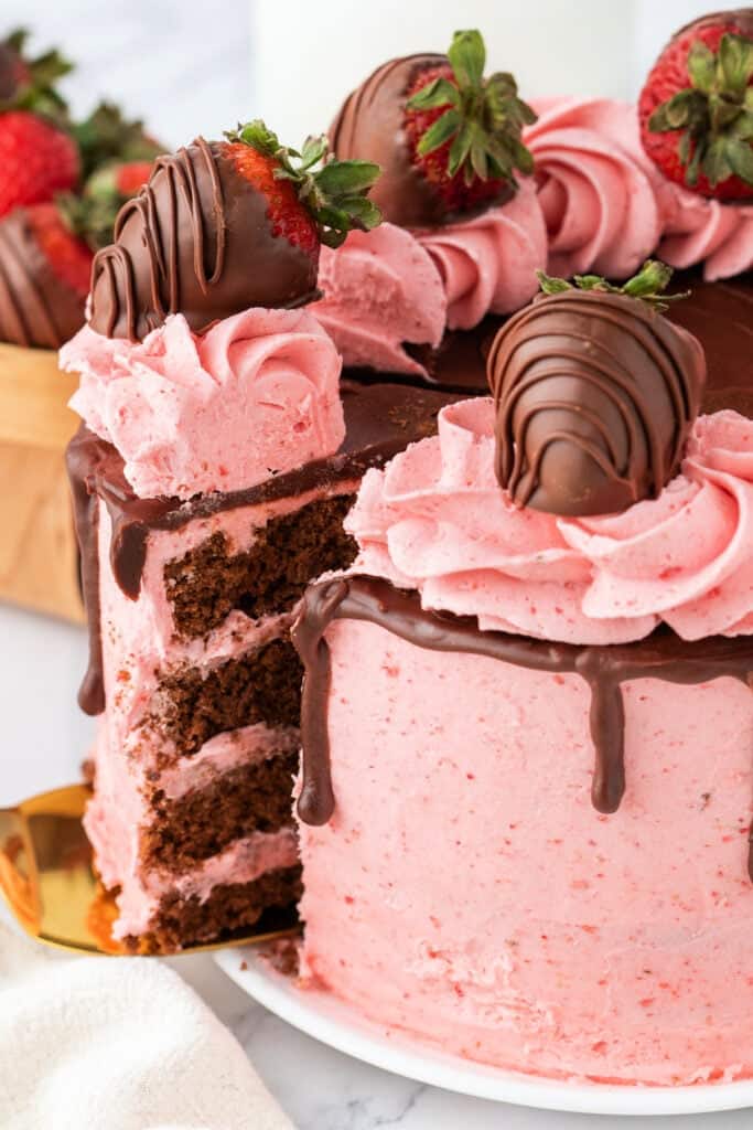 Sliced chocolate strawberry cake with pink frosting and chocolate ganache, topped with chocolate-covered strawberries.